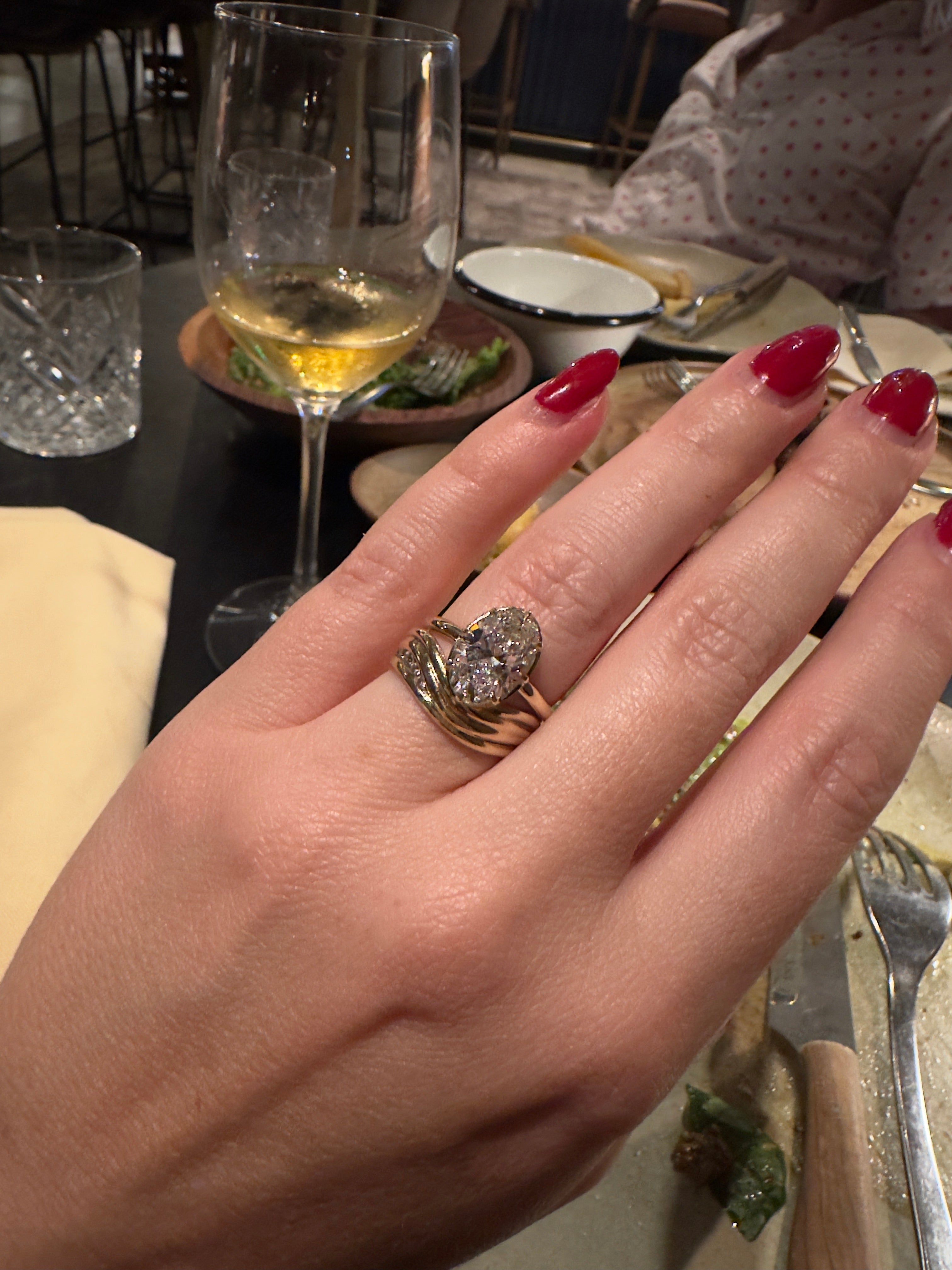 Hand with a diamond ring holding a glass of champagne, with a blurred background of a dining setting.
