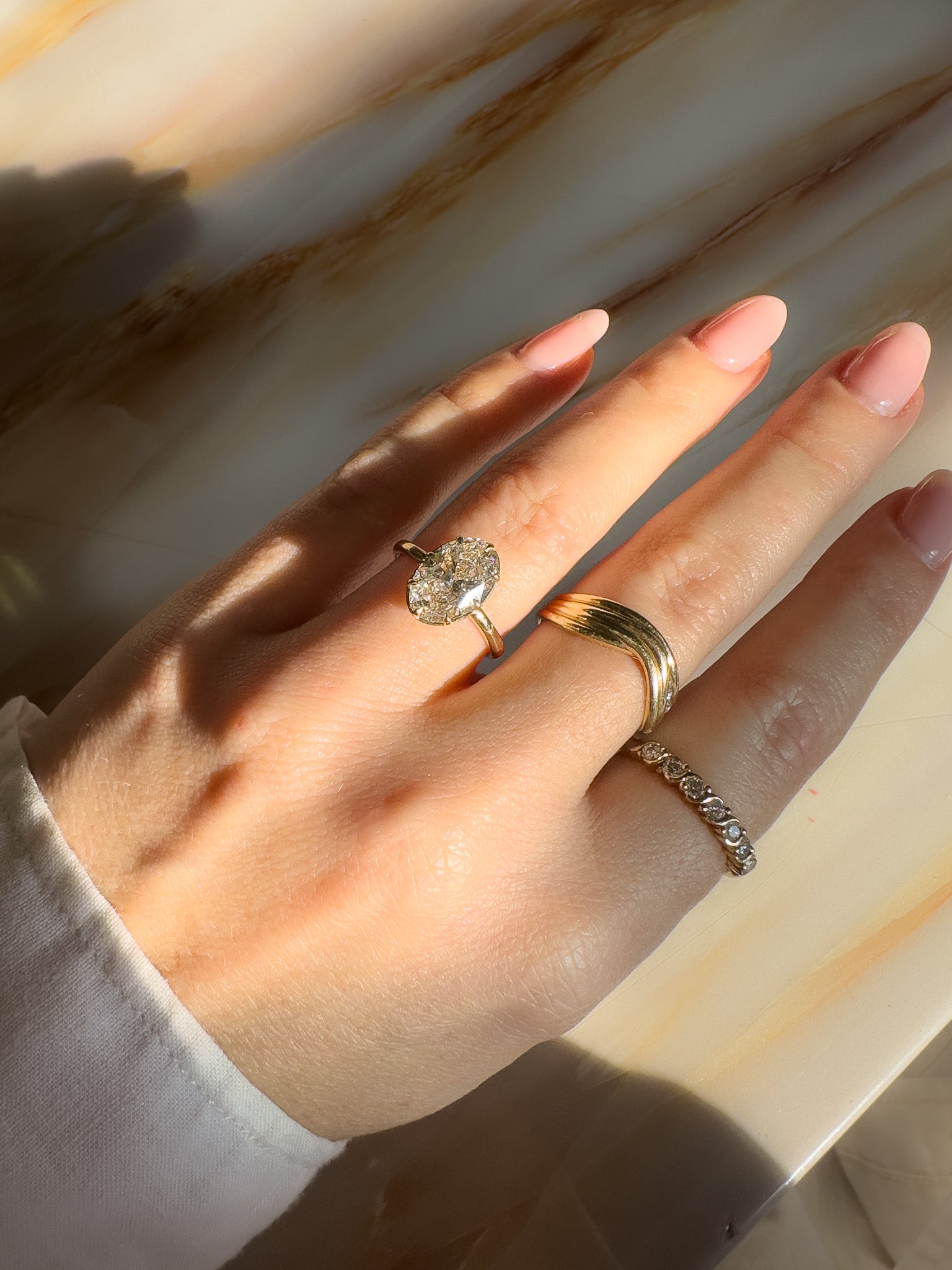 A close-up of a hand wearing a gold engagement ring with a large 3ct diamond, along with two gold bands, set against a warm marble background with strong sunlight and shadows.