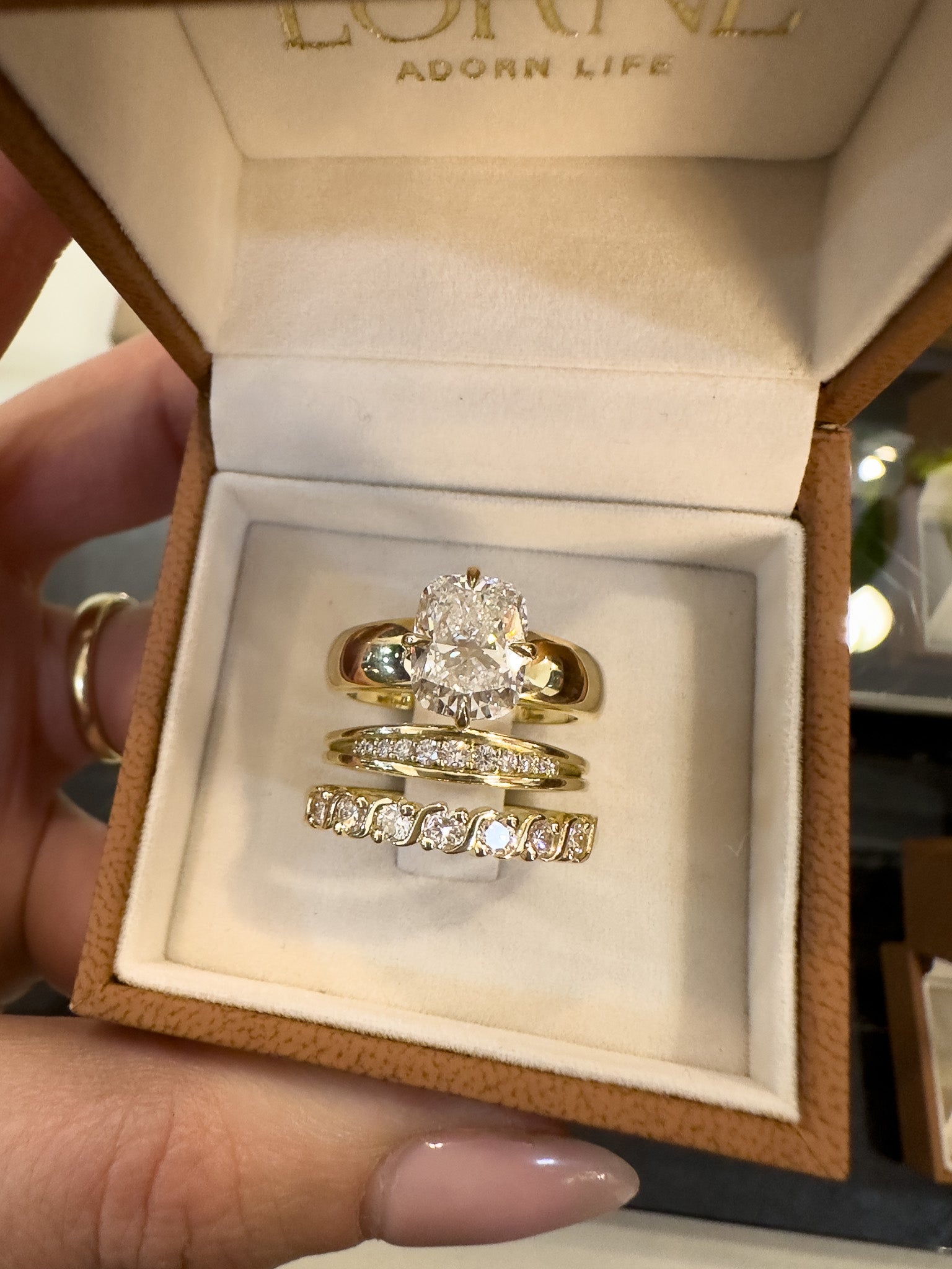 A close-up of a hand holding an open brown jewelry box with a cream velvet insert, displaying three gold rings with diamond and moisgemstones. The top ring features a large elongated cushion cut center stone, while the middle and bottom rings are gold bands with rows of small clear stones.