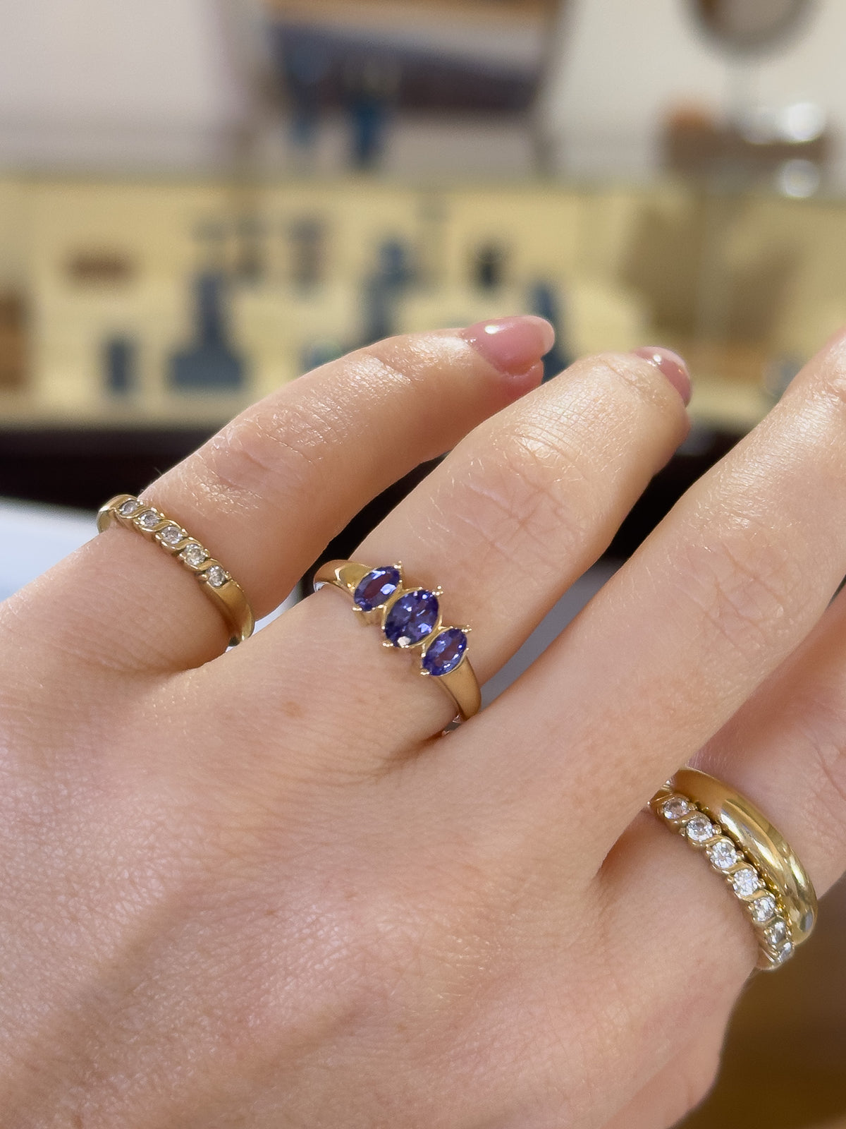 A close-up of a hand wearing multiple gold rings, including a central ring with three oval purple gemstones and two diamond-set rings on other fingers.