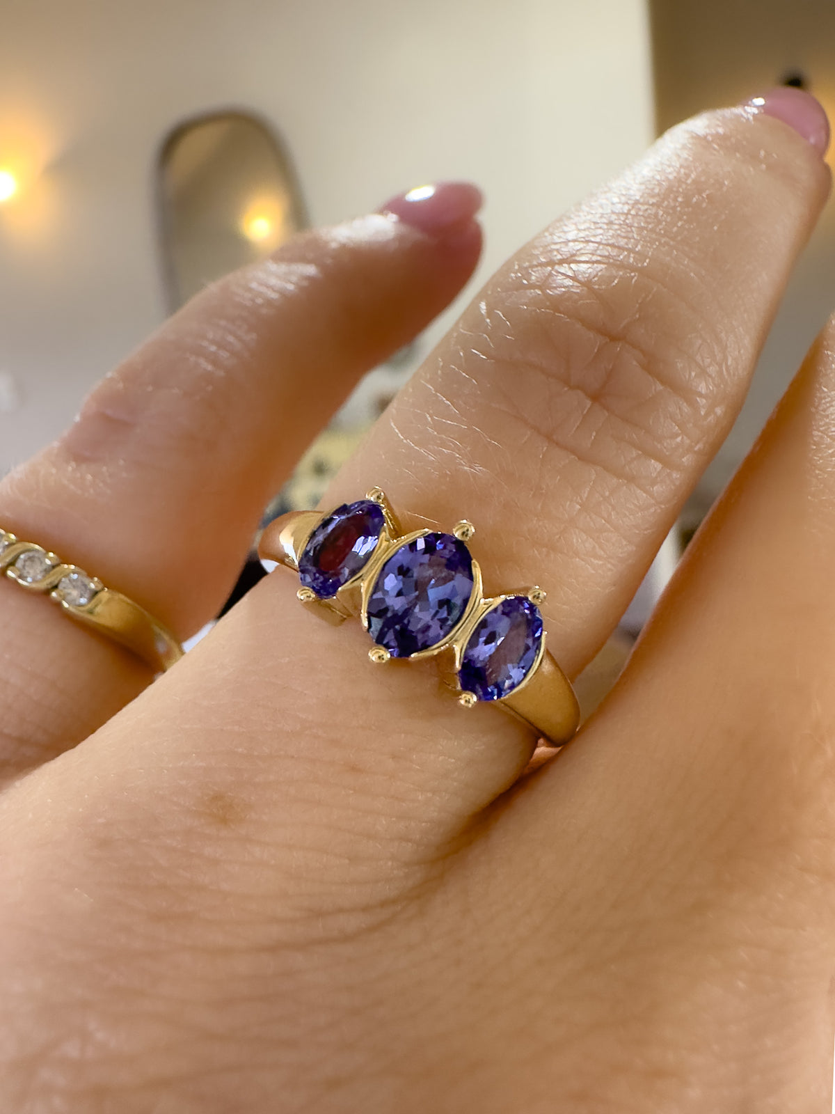 A gold ring with three oval-cut purple tanzanite gemstones worn on a finger, shown in a close-up lifestyle shot.