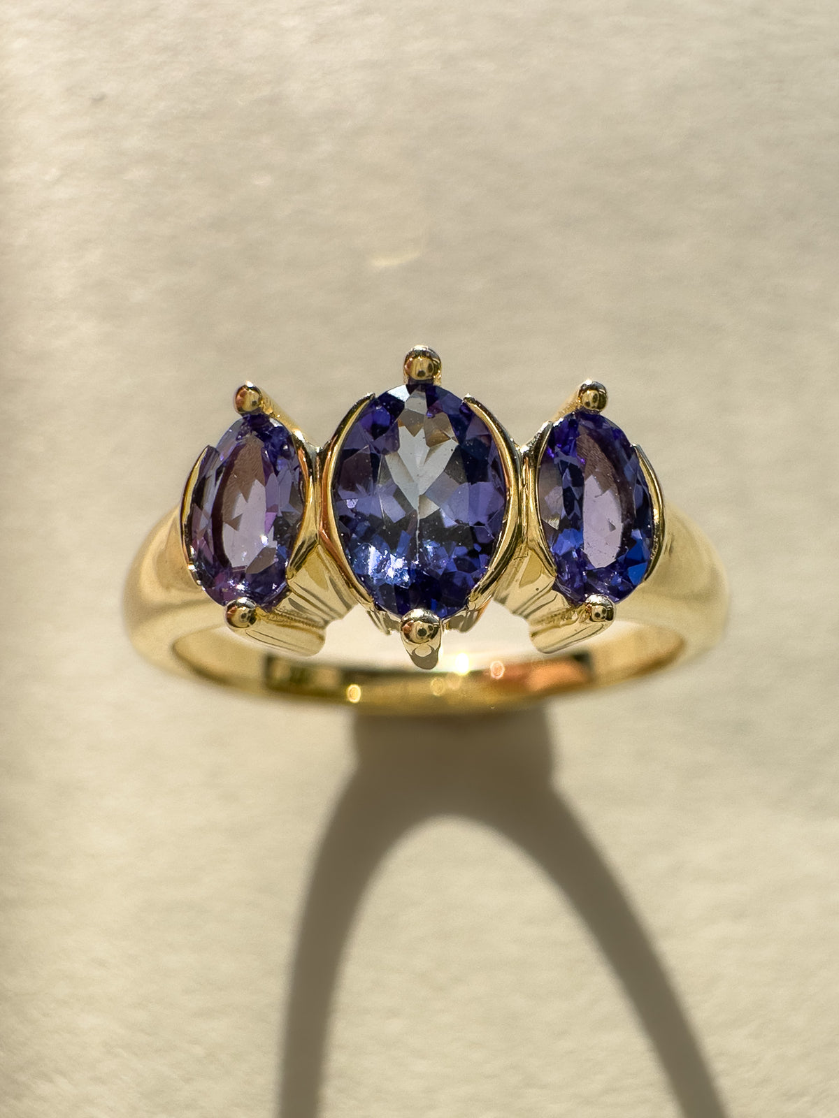A yellow gold ring featuring three oval-cut tanzanite gemstones in a row, displayed on a textured beige surface with a soft shadow.