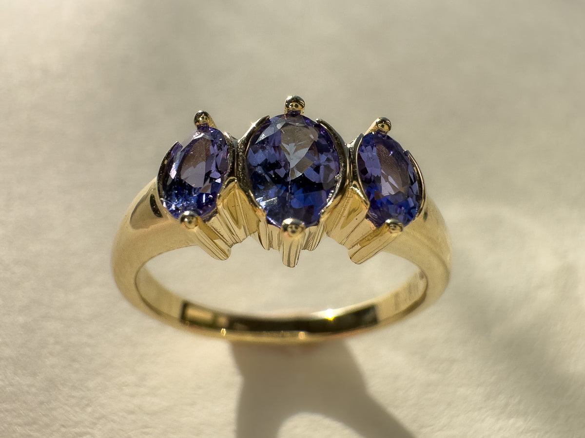 A yellow gold ring featuring three oval-cut deep blue gemstones in a prong setting, displayed on a textured light beige surface.