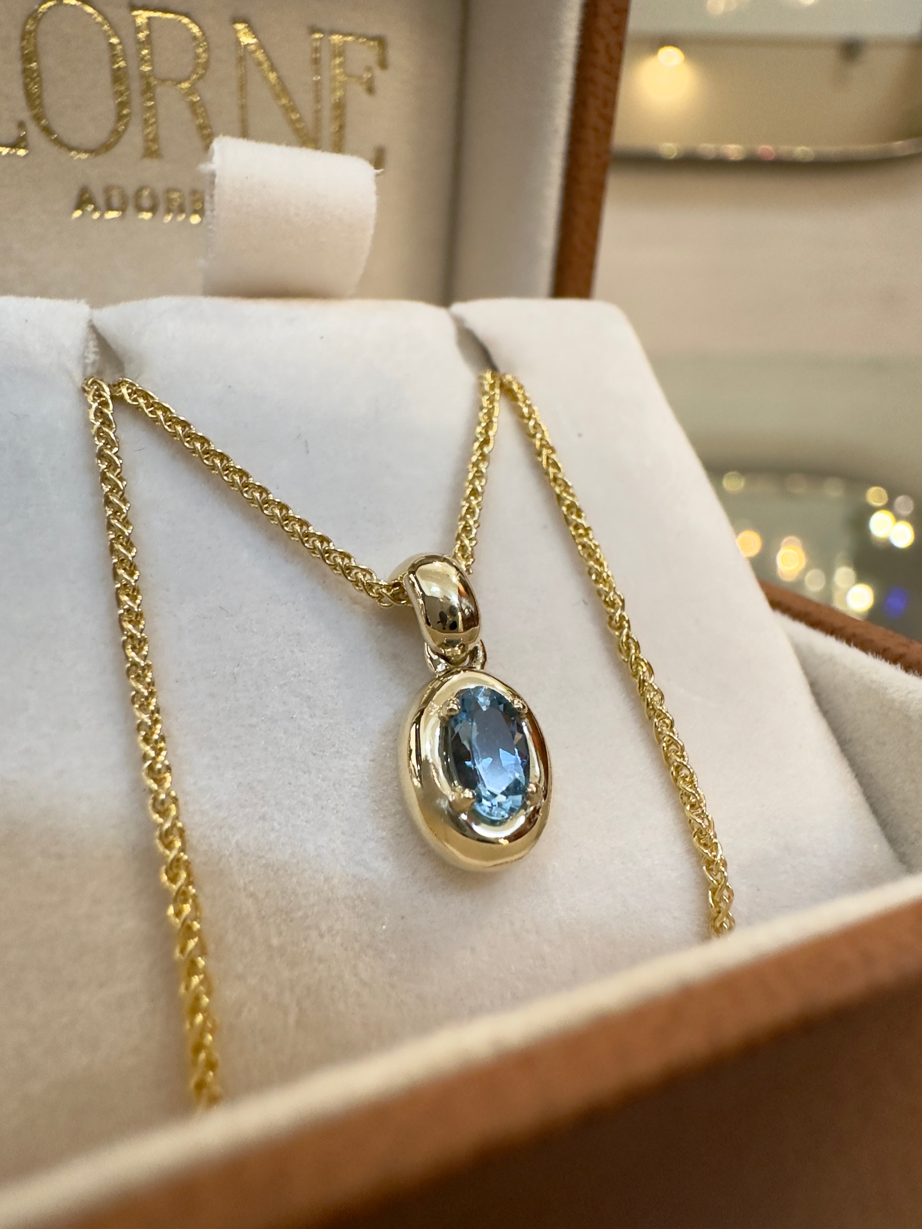 Gold necklace with a blue gemstone pendant on a white cushion, brand name visible in the background.