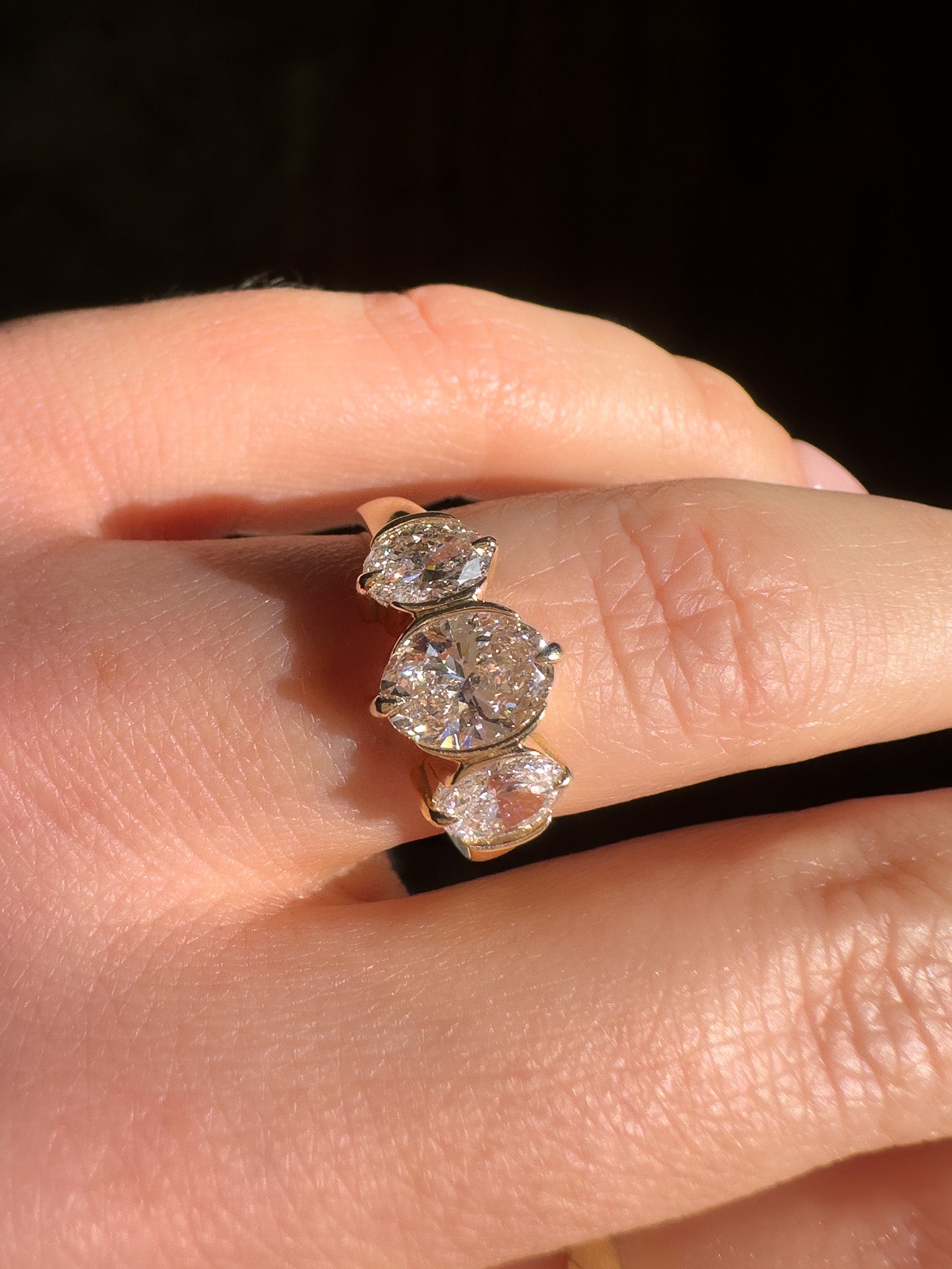 A close-up of a hand wearing a yellow gold ring with three oval-cut clear gemstones, set against a dark background.