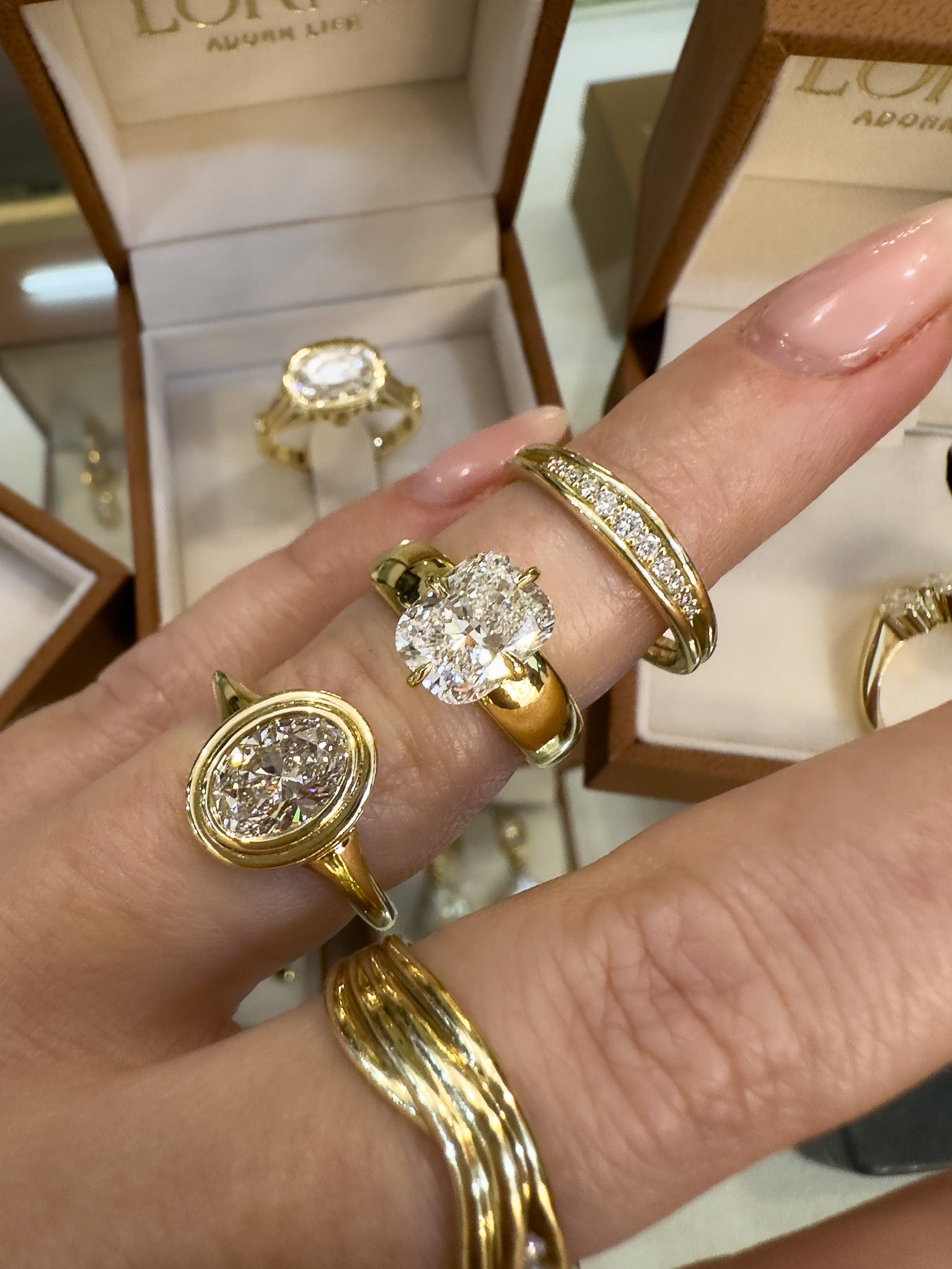 A close-up of a hand wearing multiple gold rings with diamonds, with open  jewelry boxes visible in the background.