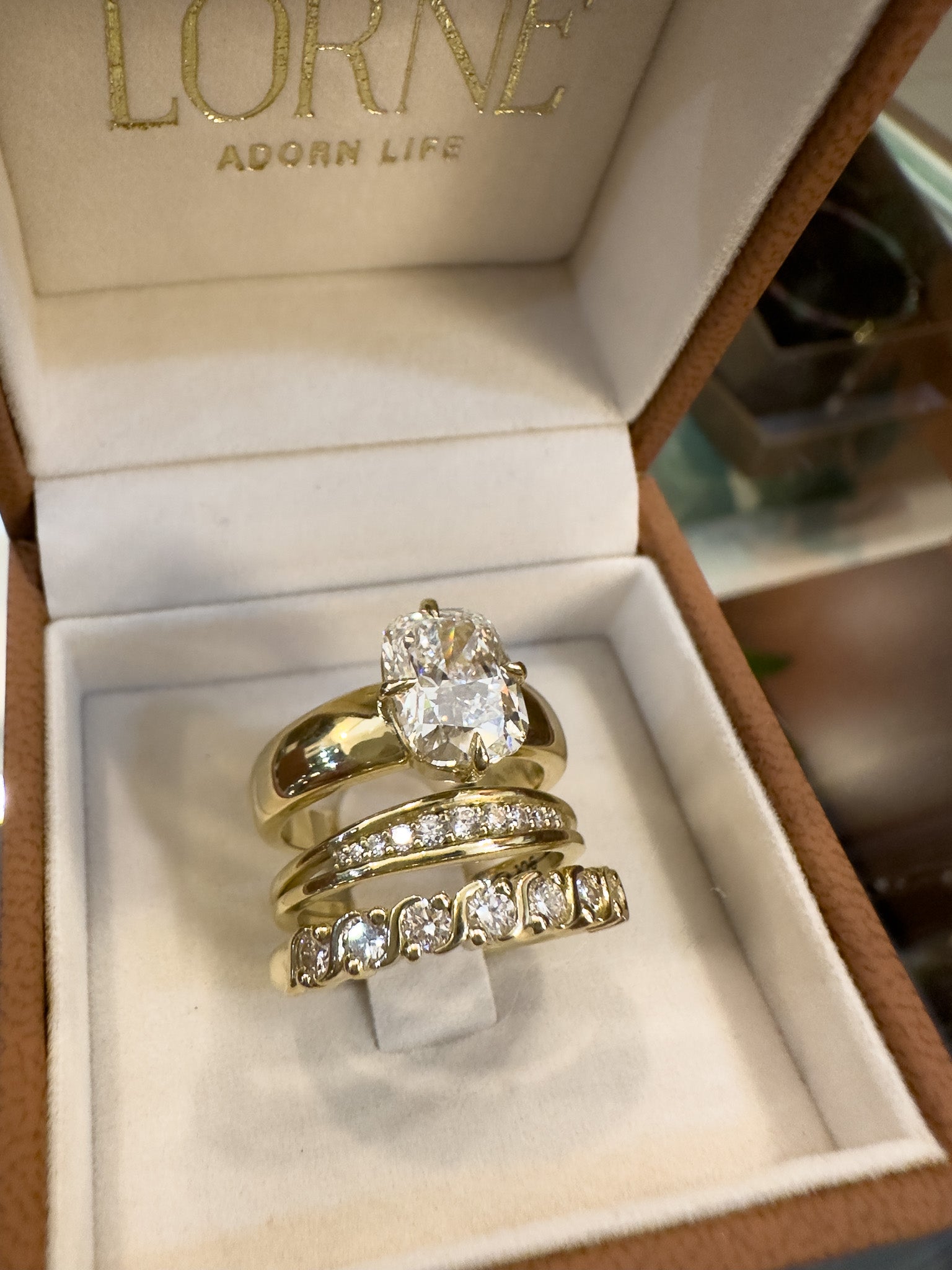 A stack of three yellow gold rings with clear gemstones displayed inside an open white jewelry box. The brand name 'LOVE ADOORN LIFE' is printed inside the lid.