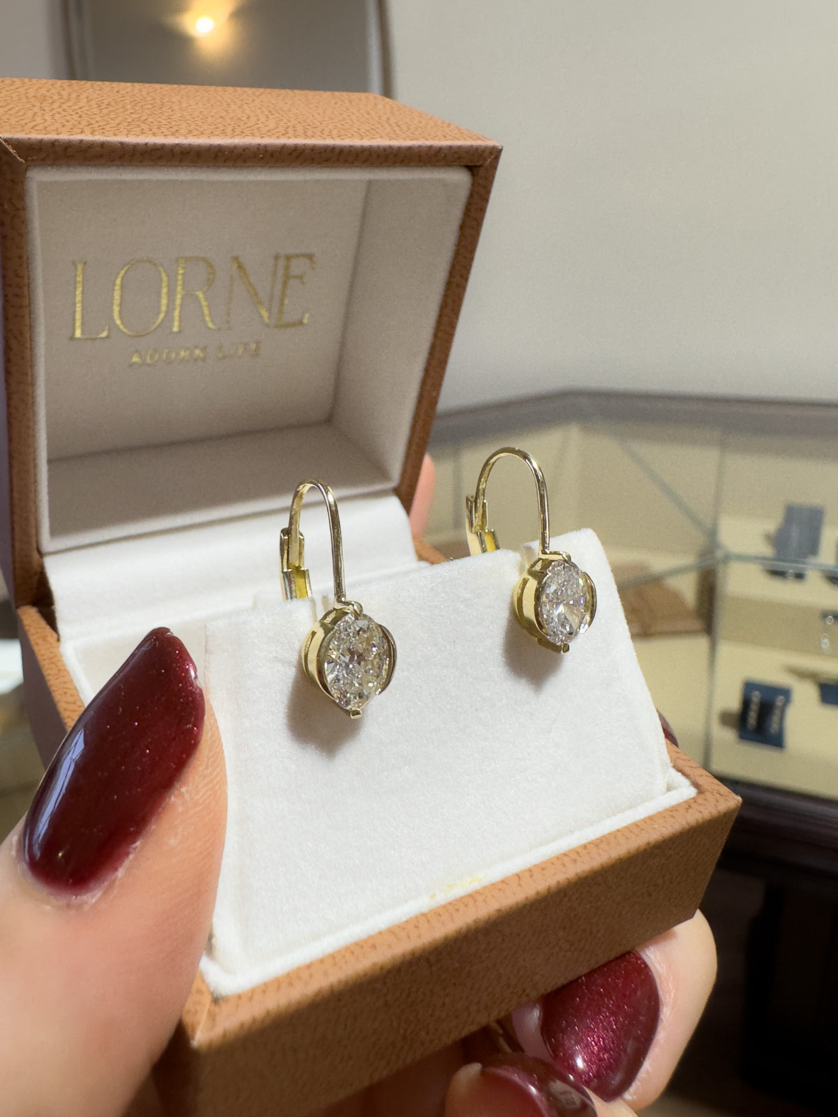 A pair of gold hoop earrings with oval clear gemstones displayed in an open LORNE jewelry box, held by a hand with dark red manicured nails.