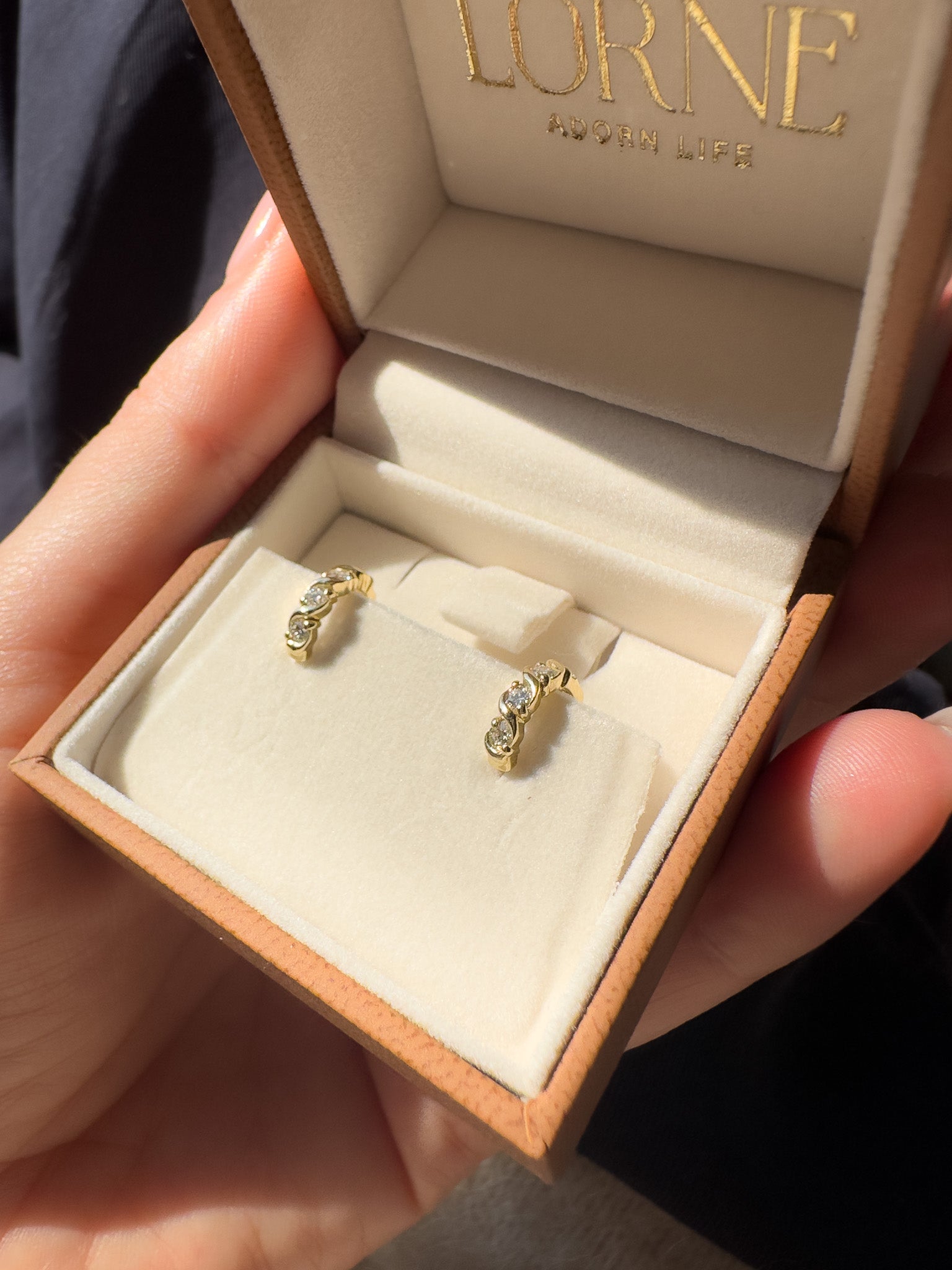 A close-up of a hand holding an open LORNE jewelry box. Inside, a pair of small gold hoop earrings with clear stones is displayed on a cream-colored velvet cushion.