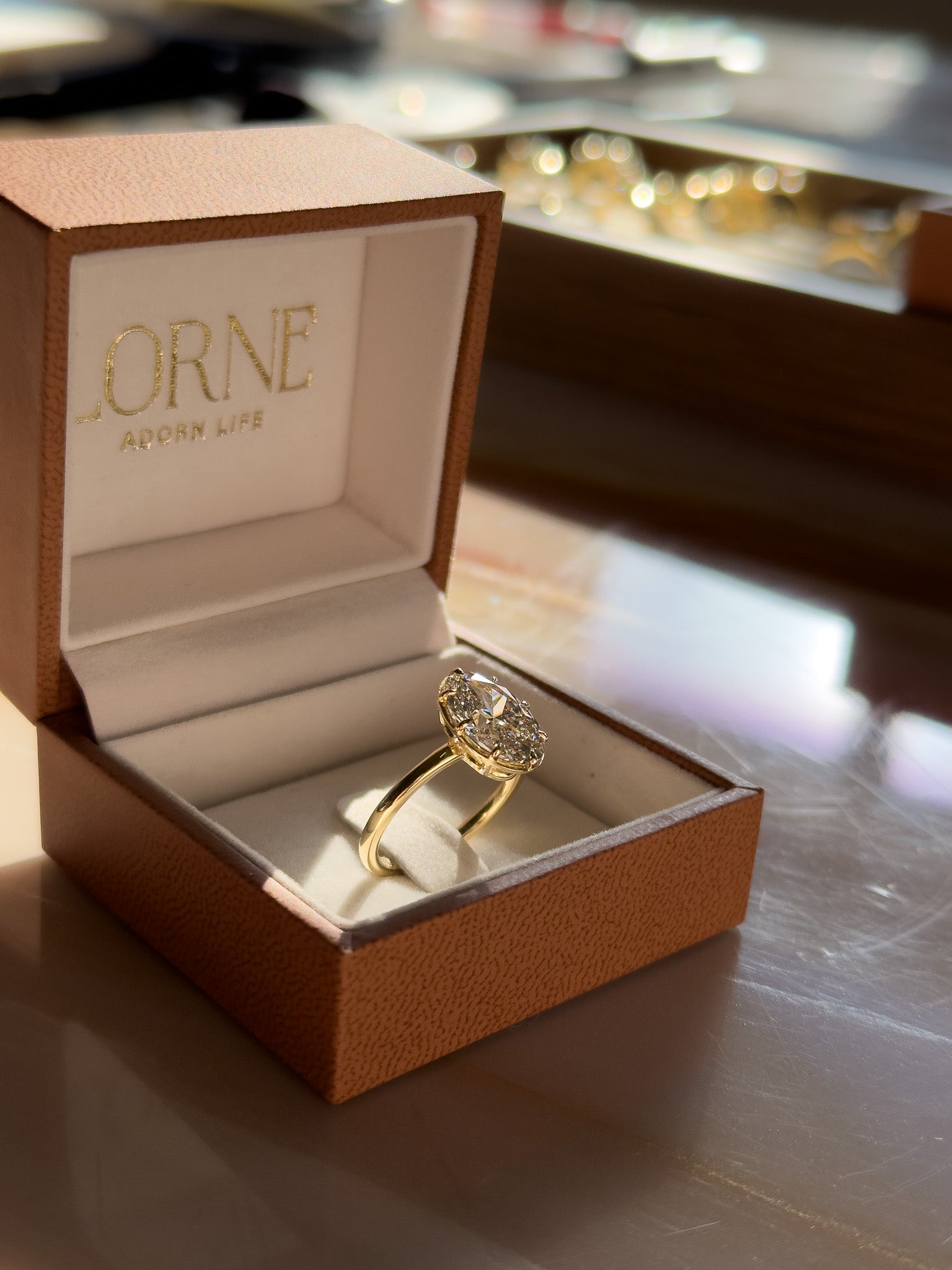 A gold ring with a large round diamond displayed in an open brown textured jewelry box. The inside lid is lined with white fabric and features the brand name 'LORNE ADON LIFE' in gold lettering.