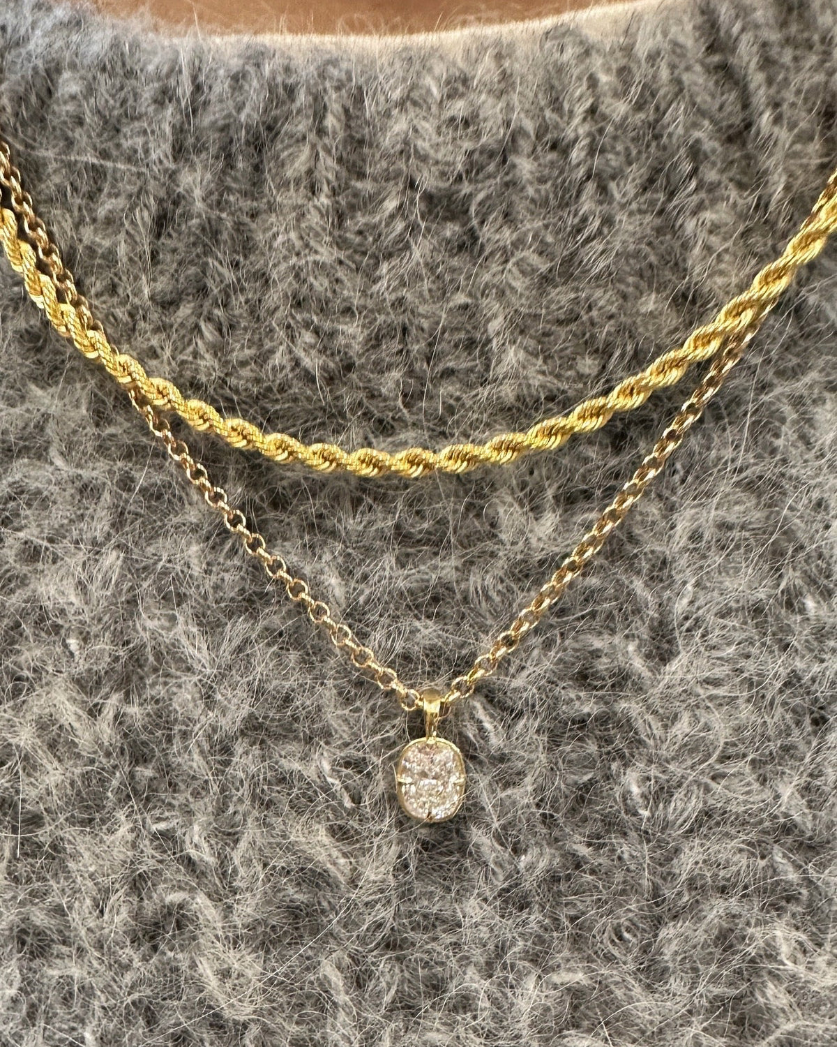 A close-up of a person wearing a layered necklace set over a gray textured knit sweater. The set includes a thicker twisted gold chain and a delicate gold chain with a small oval clear gemstone pendant.
