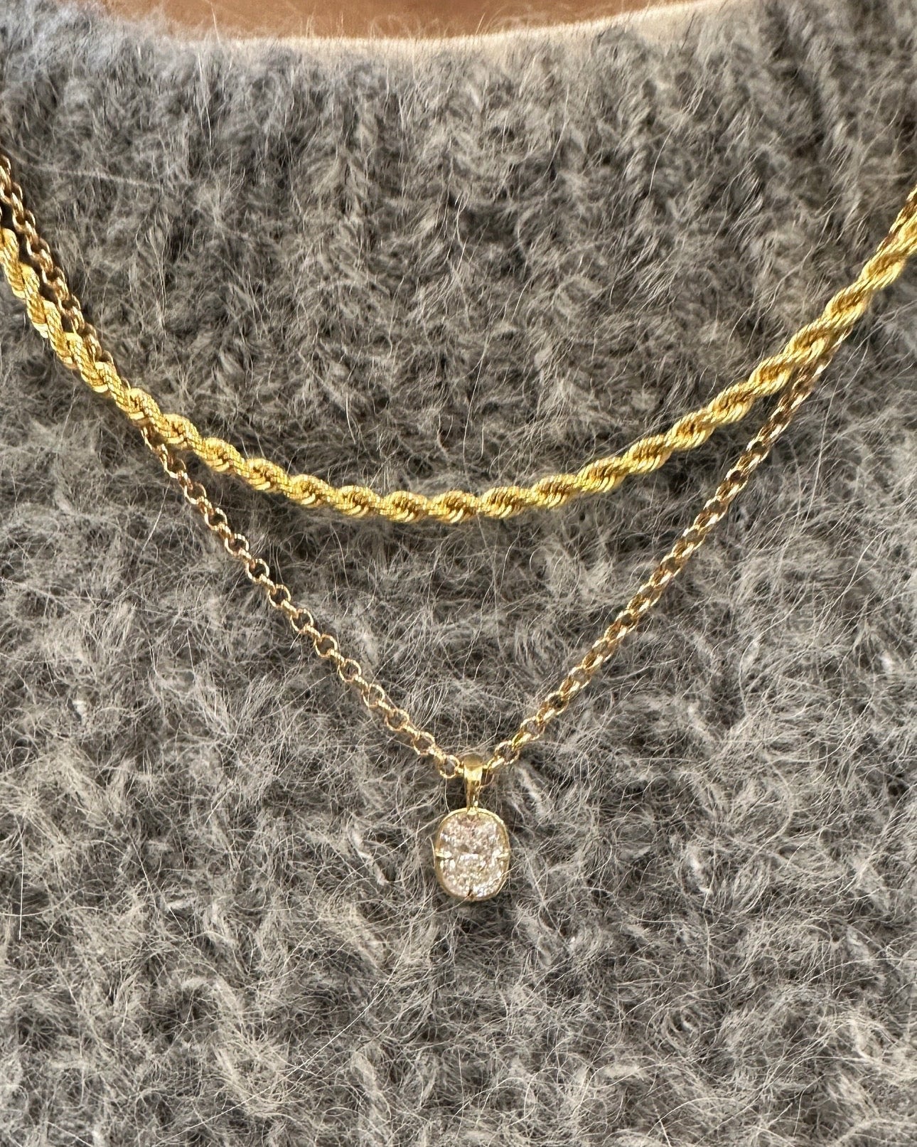 A close-up of a person wearing a layered necklace set over a gray textured knit sweater. The set includes a thicker twisted gold chain and a delicate gold chain with a small oval clear gemstone pendant.