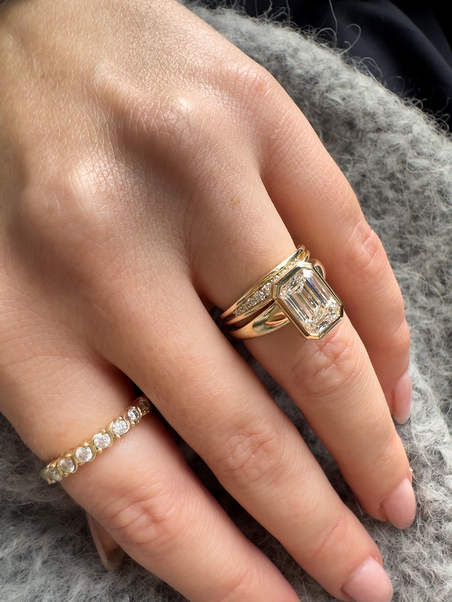 A close-up of a hand wearing a gold ring with a large emerald-cut diamond center stone, alongside a smaller diamond band ring, resting on a gray textured fabric.