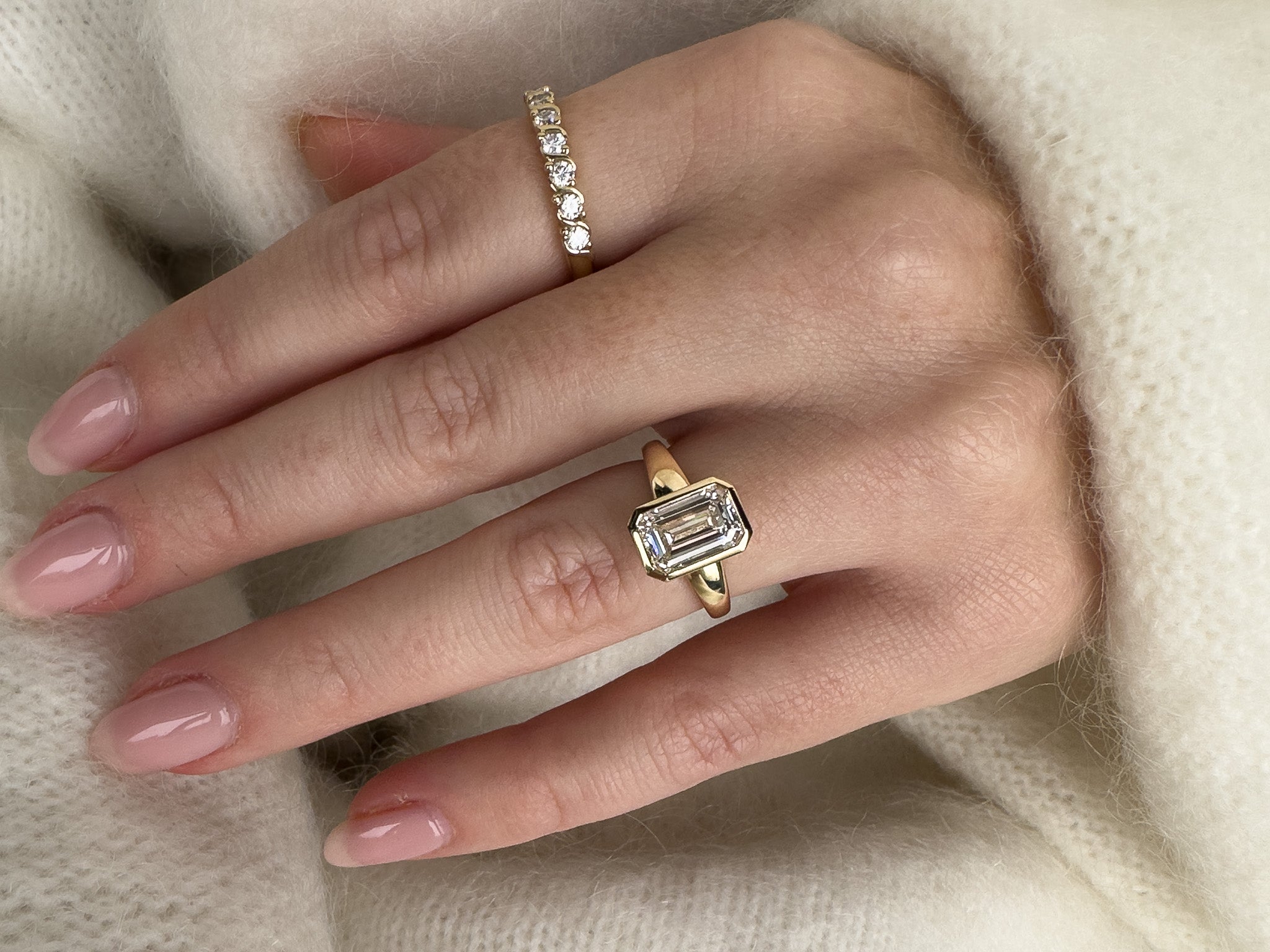 A close-up of a hand wearing two gold rings against a soft cream knit background. The larger ring on the middle finger features a rectangular emerald-cut clear gemstone, while the smaller ring on the ring finger has a row of small clear stones.