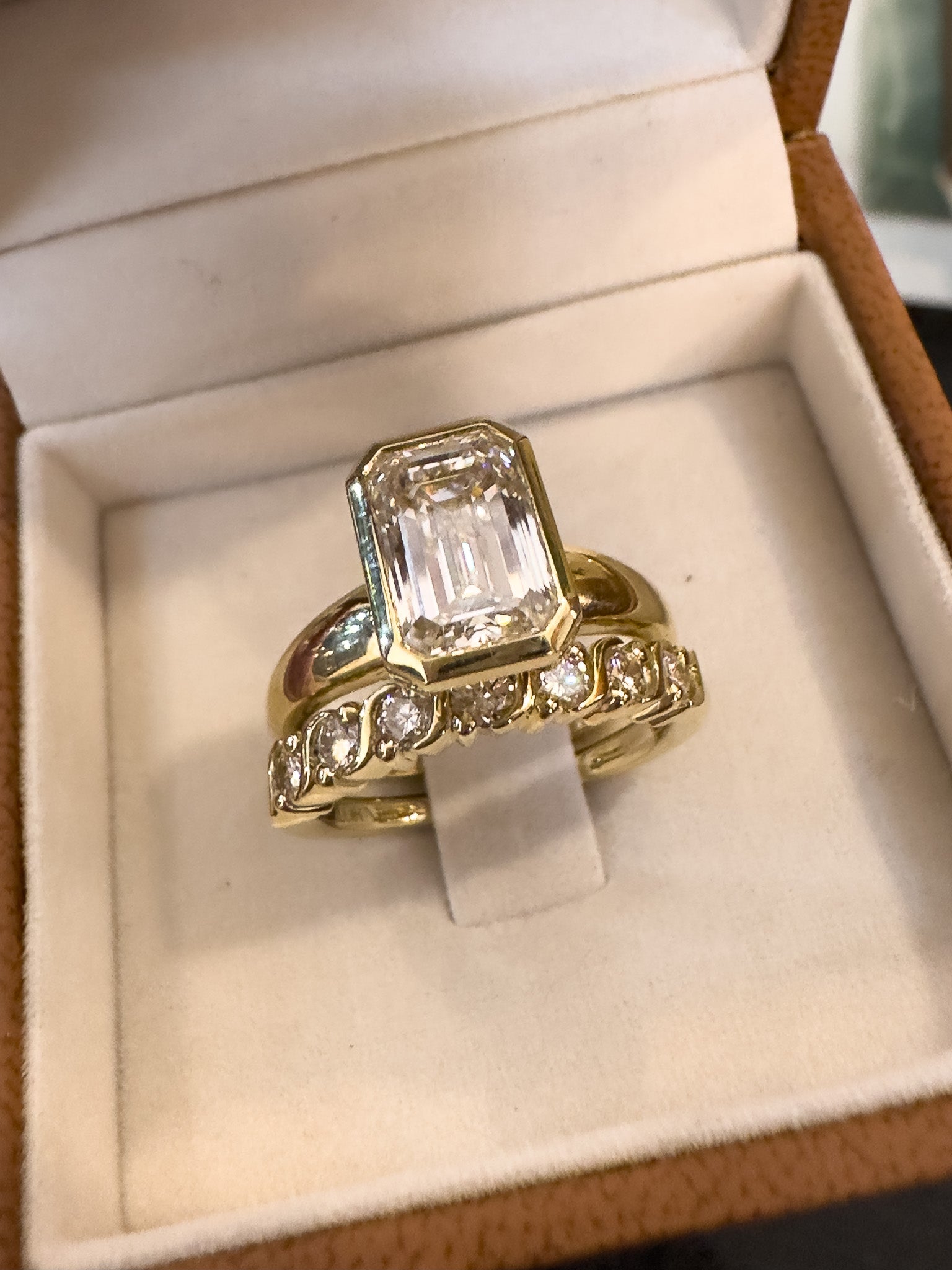 A yellow gold ring set featuring a large emerald-cut clear center stone and smaller accent stones along the band, displayed inside an open cream-colored jewelry box.
