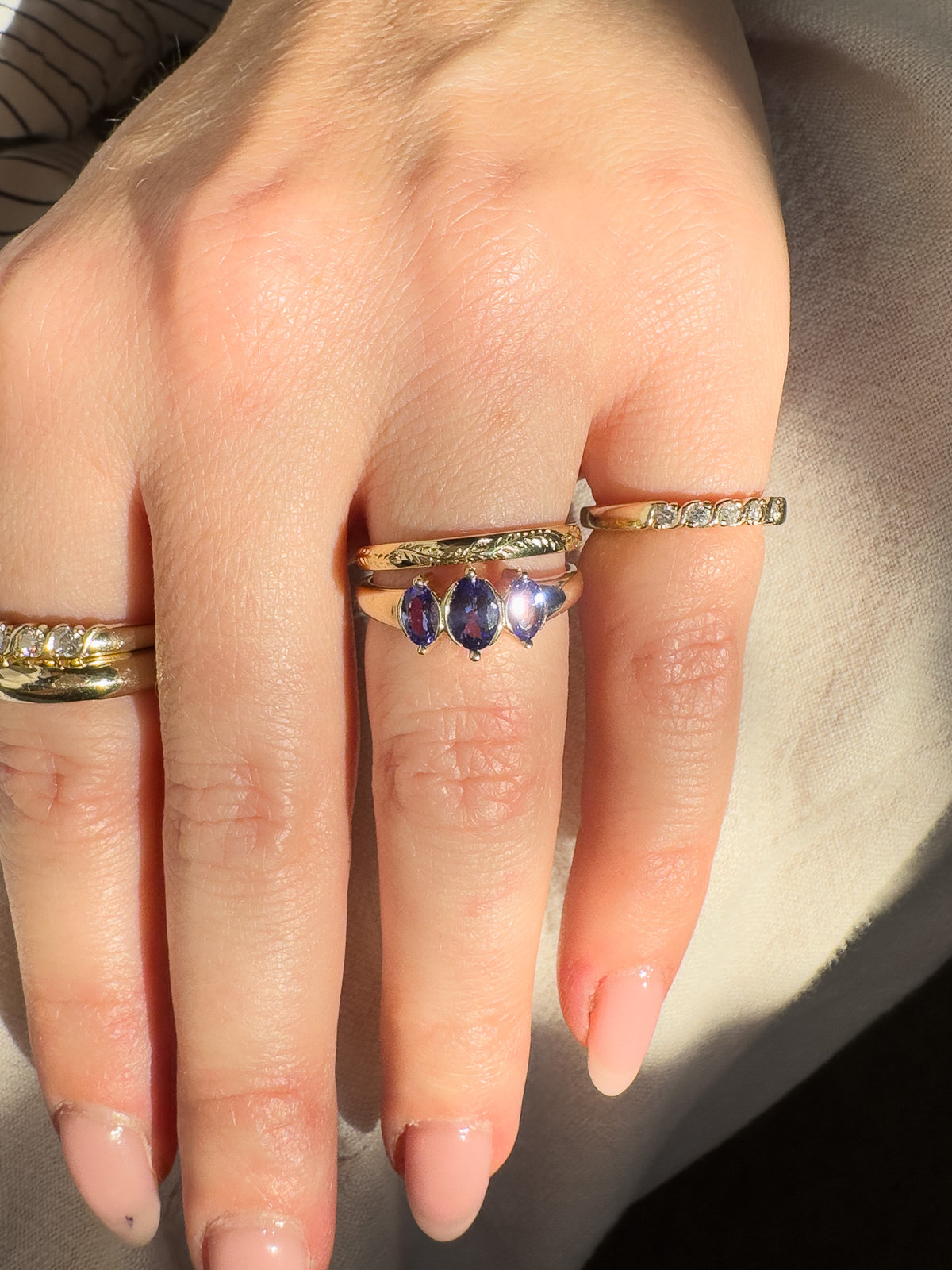 A close-up of a hand wearing multiple rings, with the centre finger featuring a gold band with three marquise-cut blue-purple gemstones, flanked by gold bands with clear stones. South Africa.
