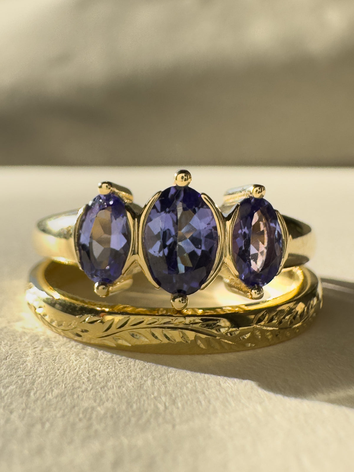 A gold ring set with three oval-cut deep blue-purple tanzanite gemstones, displayed on top of a matching engraved gold band against a neutral beige background.