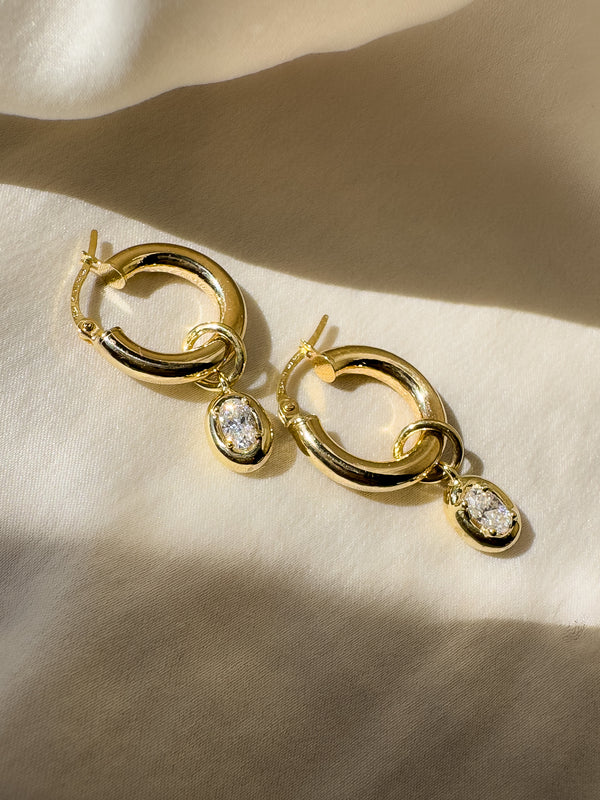 Gold hoop earrings with teardrop-shaped gemstones on a beige fabric background