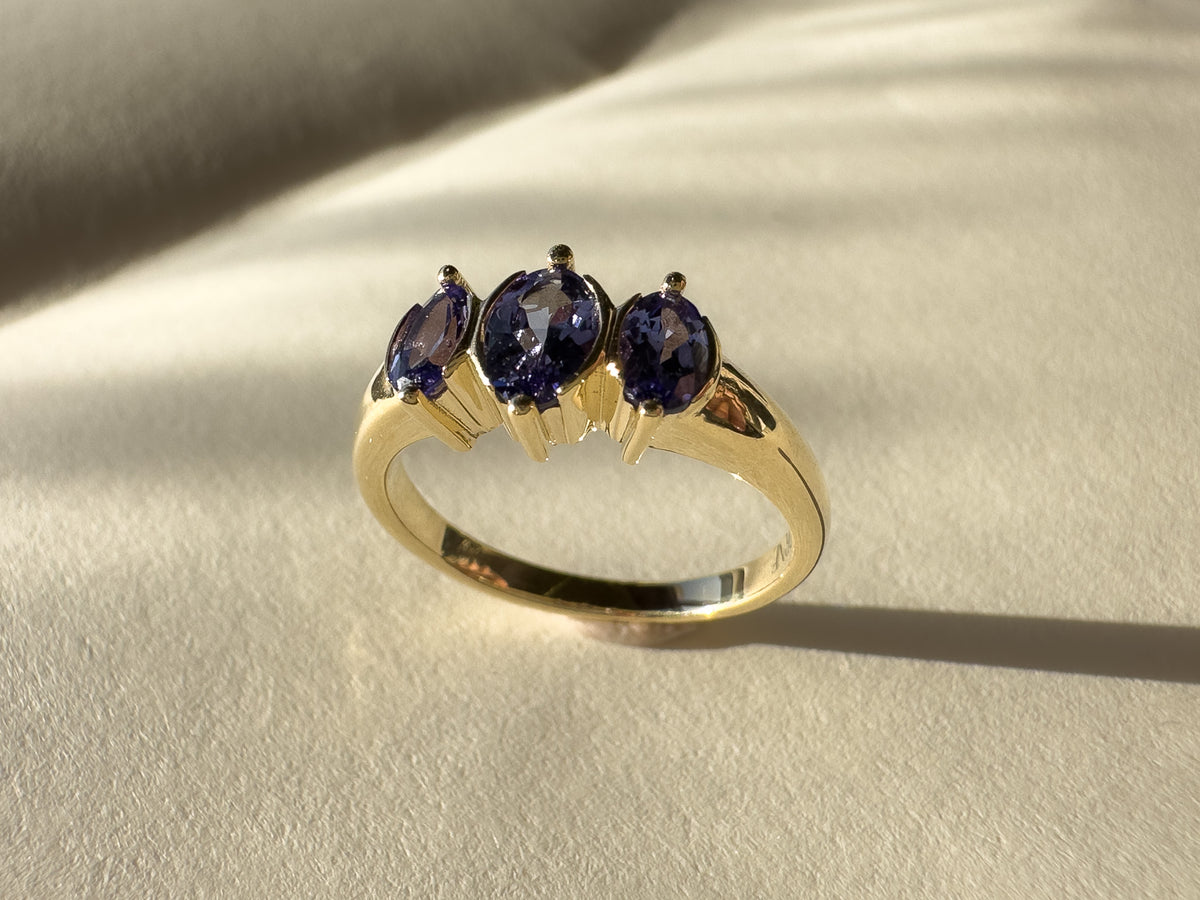 A yellow gold ring with three oval-cut dark blue gemstones, displayed on a textured beige surface with soft shadows.