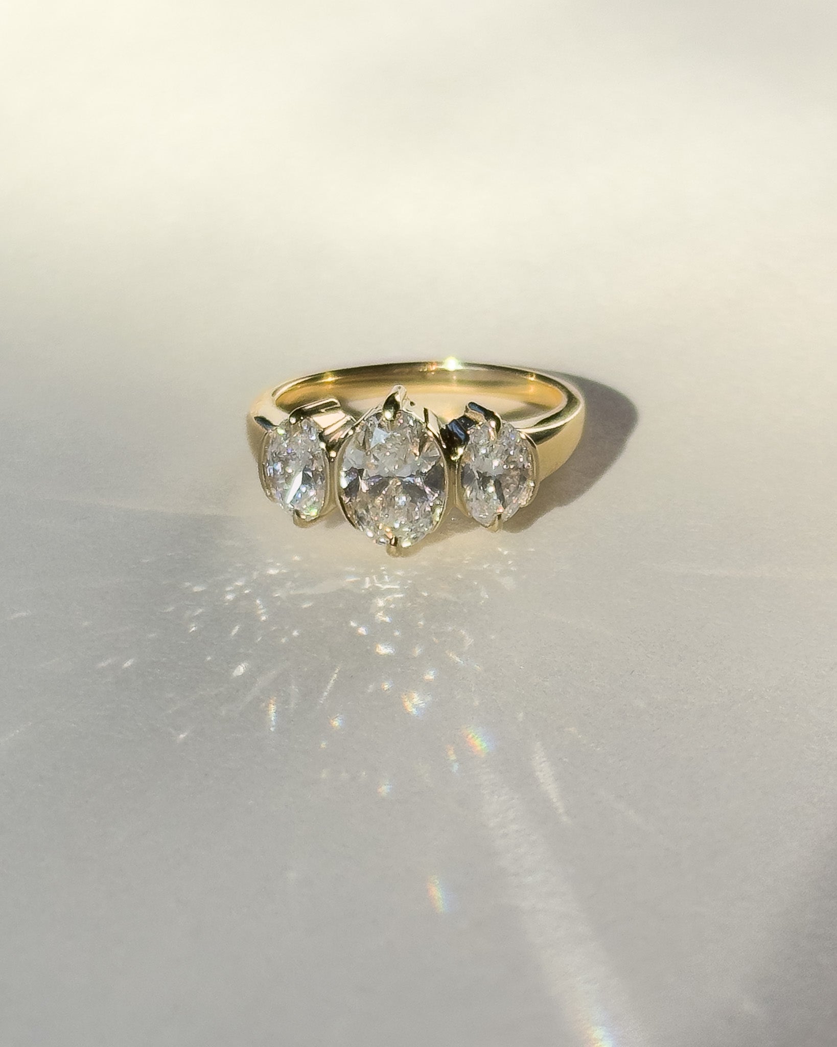 A yellow gold ring with three oval-cut clear gemstones, photographed on a light gray surface with soft shadows and light reflections.