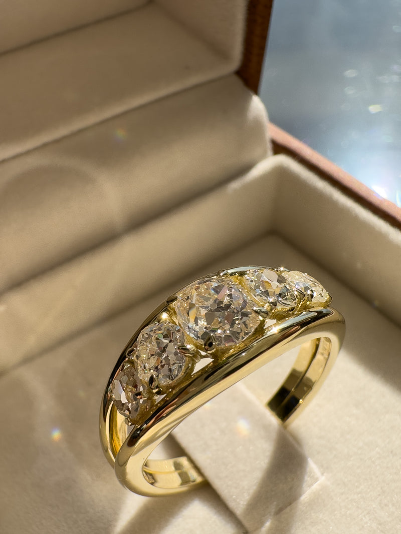 A yellow gold ring with five round-cut clear gemstones set in a curved row, displayed inside an open beige jewelry box.