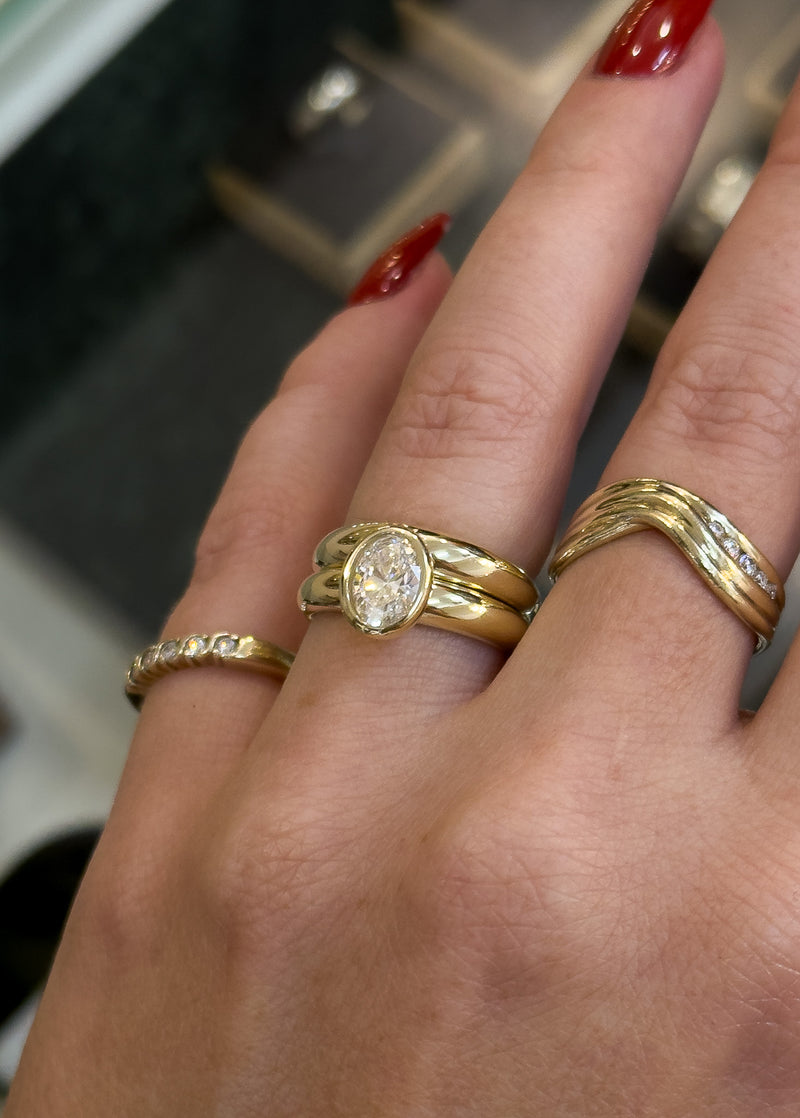 A close-up of a hand wearing multiple gold rings, including a yellow gold engagement ring with a round clear gemstone and two twisted gold bands with small clear stones.