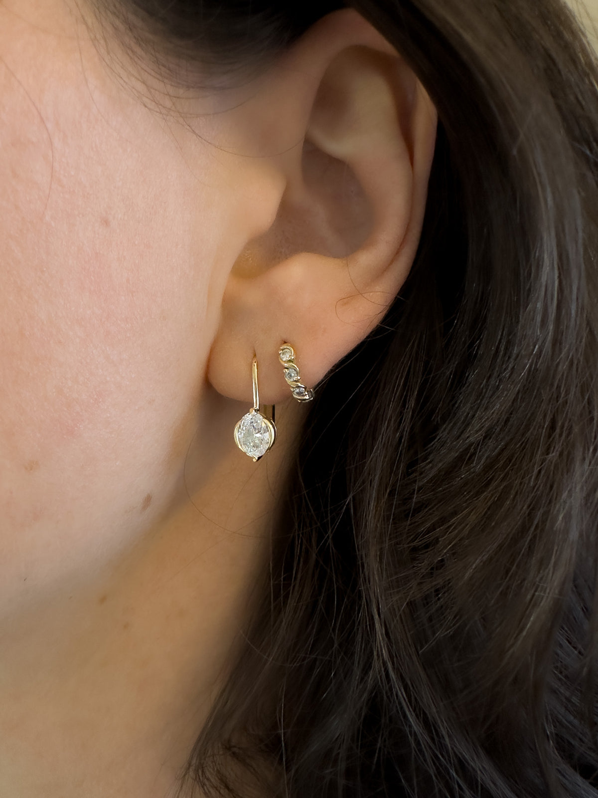 A close-up of a person's ear wearing a small gold hoop earring with a clear teardrop-shaped gemstone and a matching small gold cartilage hoop with tiny clear stones.
