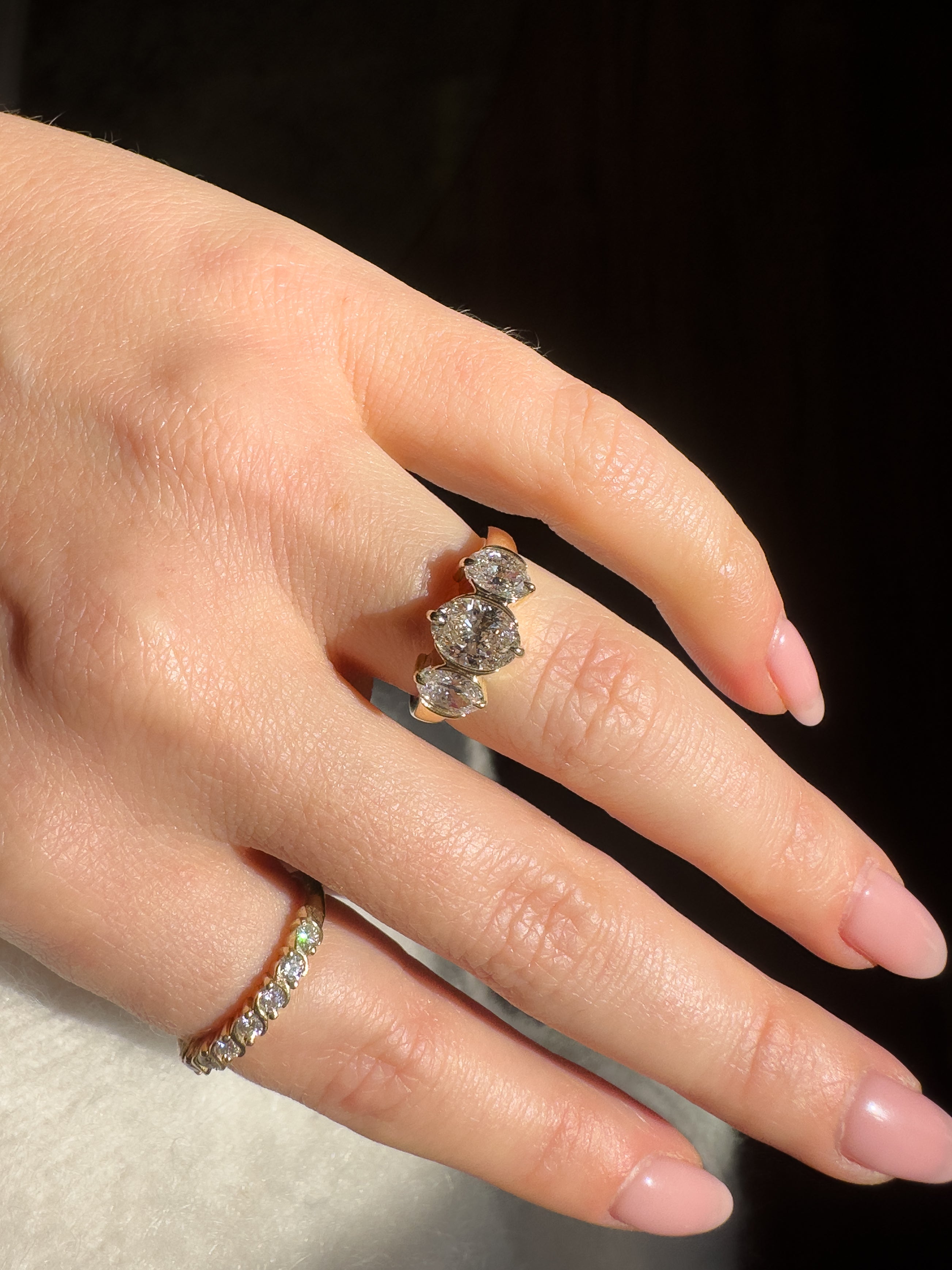 A close-up of a hand wearing a gold ring with three oval-cut clear gemstones, alongside another thin gold band ring, against a dark background.