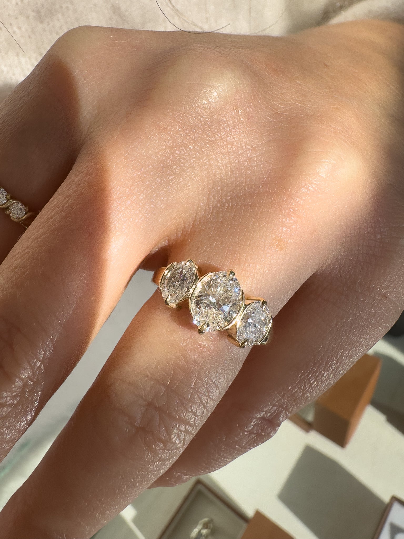A close-up of a hand wearing a yellow gold ring with three round-cut clear gemstones in a row, set against a softly blurred background.