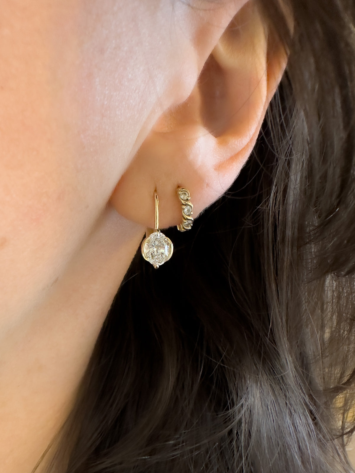 A close-up of a person's ear wearing two gold earrings: a small curved huggie hoop and a dangling drop earring with a clear round gemstone.