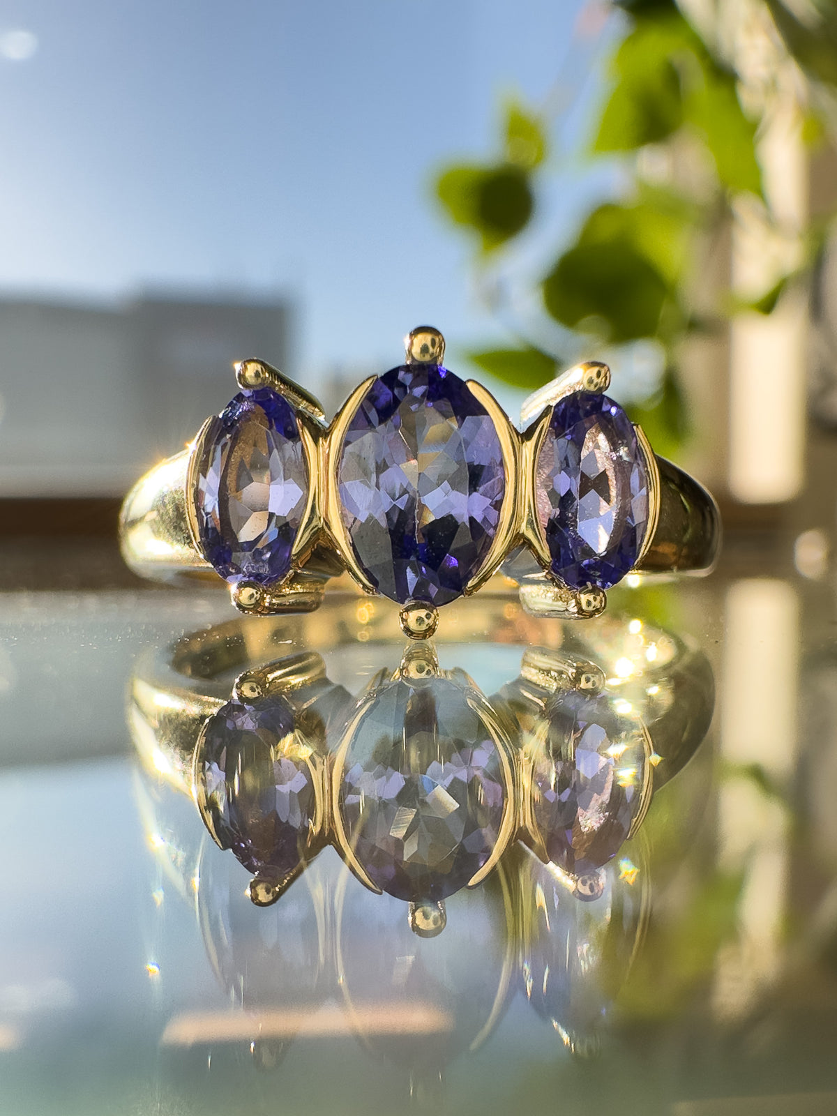 A yellow gold ring featuring three marquise-cut tanzanite gemstones, displayed on a reflective surface with a soft-focus background of blue sky and green leaves.