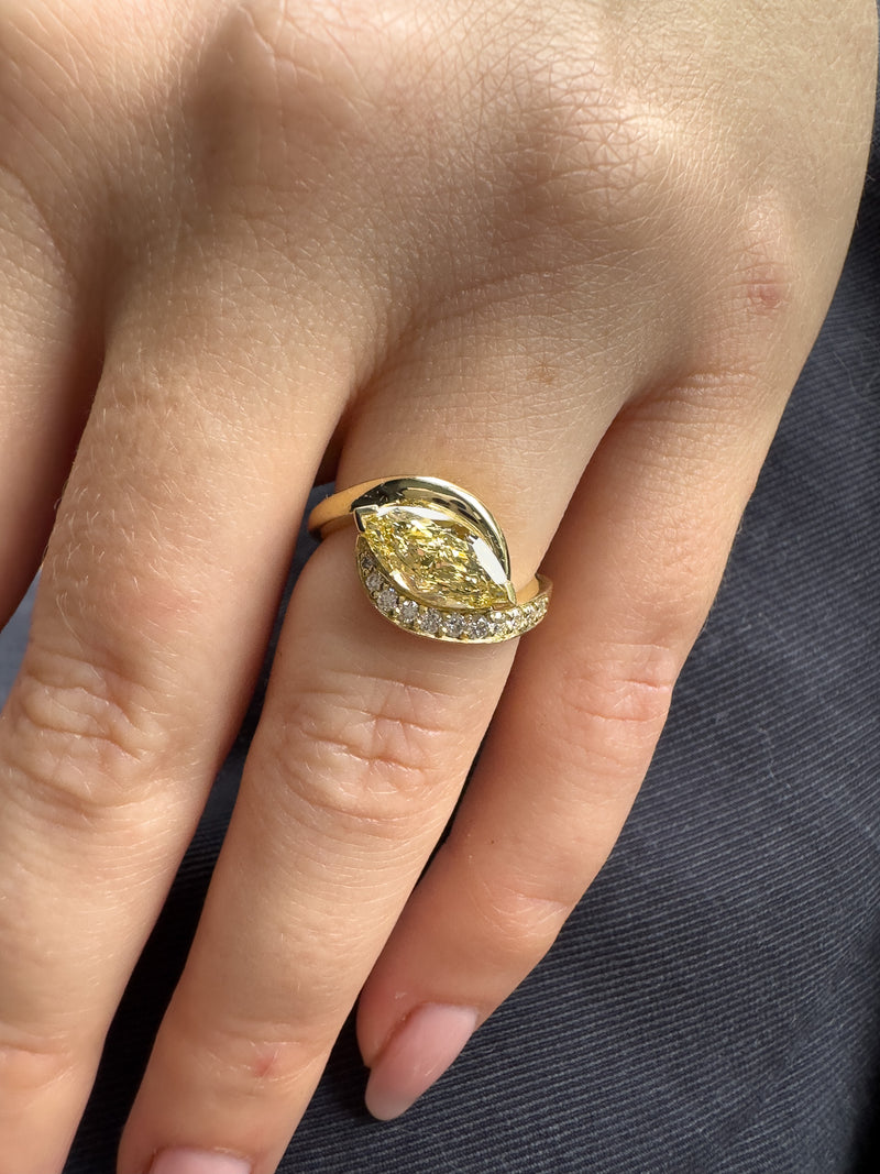 A close-up of a hand wearing a yellow gold ring with a marquise-cut yellow gemstone and a row of small clear stones along the band.